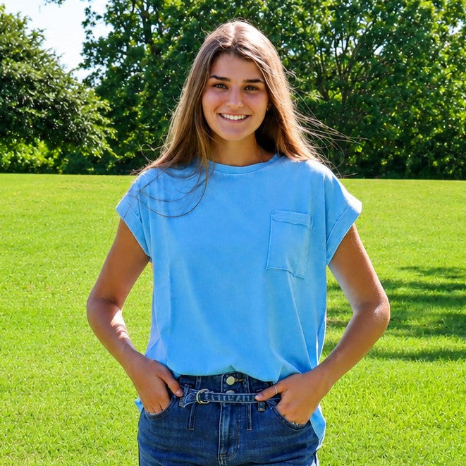 A young woman smiling outdoors in a sunny park, wearing a light blue short-sleeve t-shirt and dark blue denim shorts.