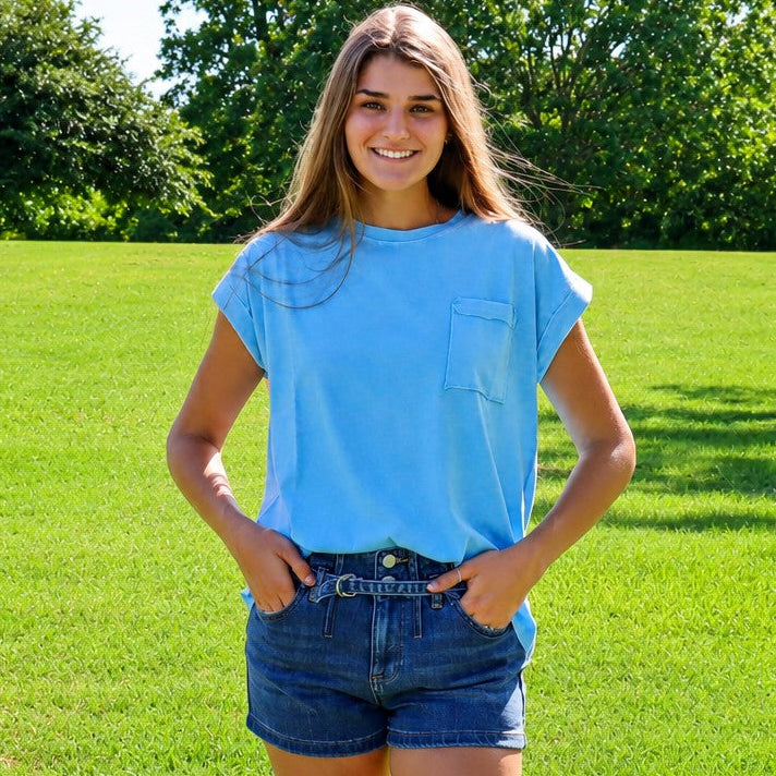 A young woman smiling outdoors in a sunny park, wearing a light blue short-sleeve t-shirt and dark blue denim shorts.