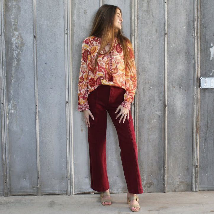 Person in a floral top and burgundy pants standing against a wooden wall.