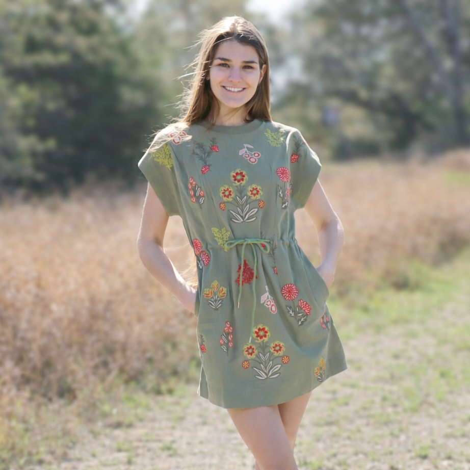 Woman wearing a green dress with floral embroidery standing in a natural setting.