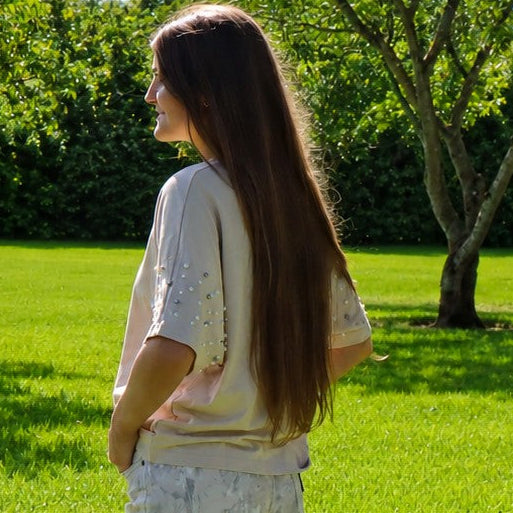 A woman with long dark hair stands on a bright green lawn in a park, wearing a light beige short-sleeve top and matching light beige wide-leg trousers with a subtle floral pattern.