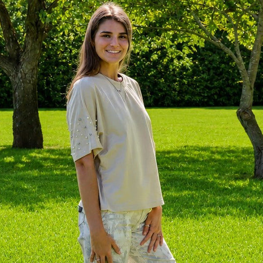 A woman stands on a bright green lawn in a park, wearing a light beige short-sleeve top and matching light-colored wide-leg pants.