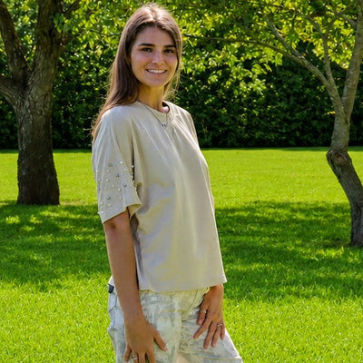 A woman stands on a bright green lawn in a park, wearing a light beige short-sleeve top and matching light-colored wide-leg pants.
