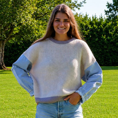 A young woman stands on a green grassy lawn wearing a light blue and off-white ribbed knit sweater with light blue denim shorts, with trees in the background.