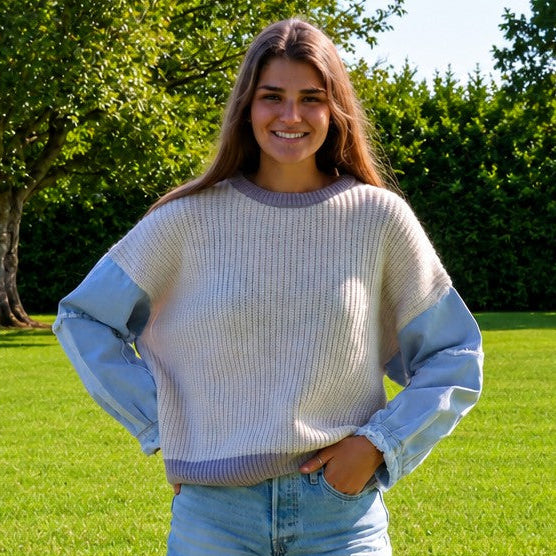 A young woman stands on a green grassy lawn wearing a light blue and off-white ribbed knit sweater with light blue denim shorts, with trees in the background.