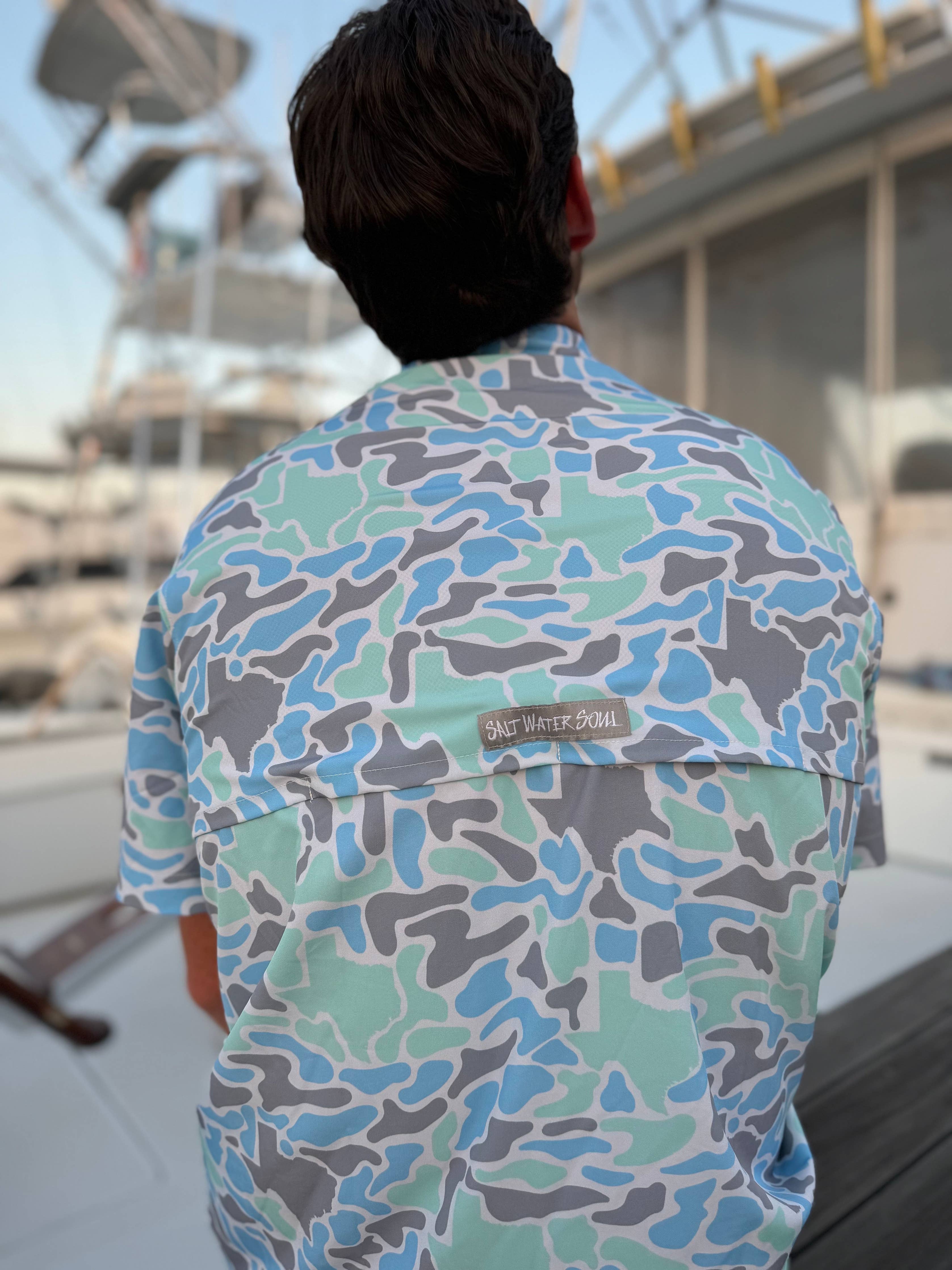 Person wearing a colorful camouflage shirt with a brand logo, standing outdoors.