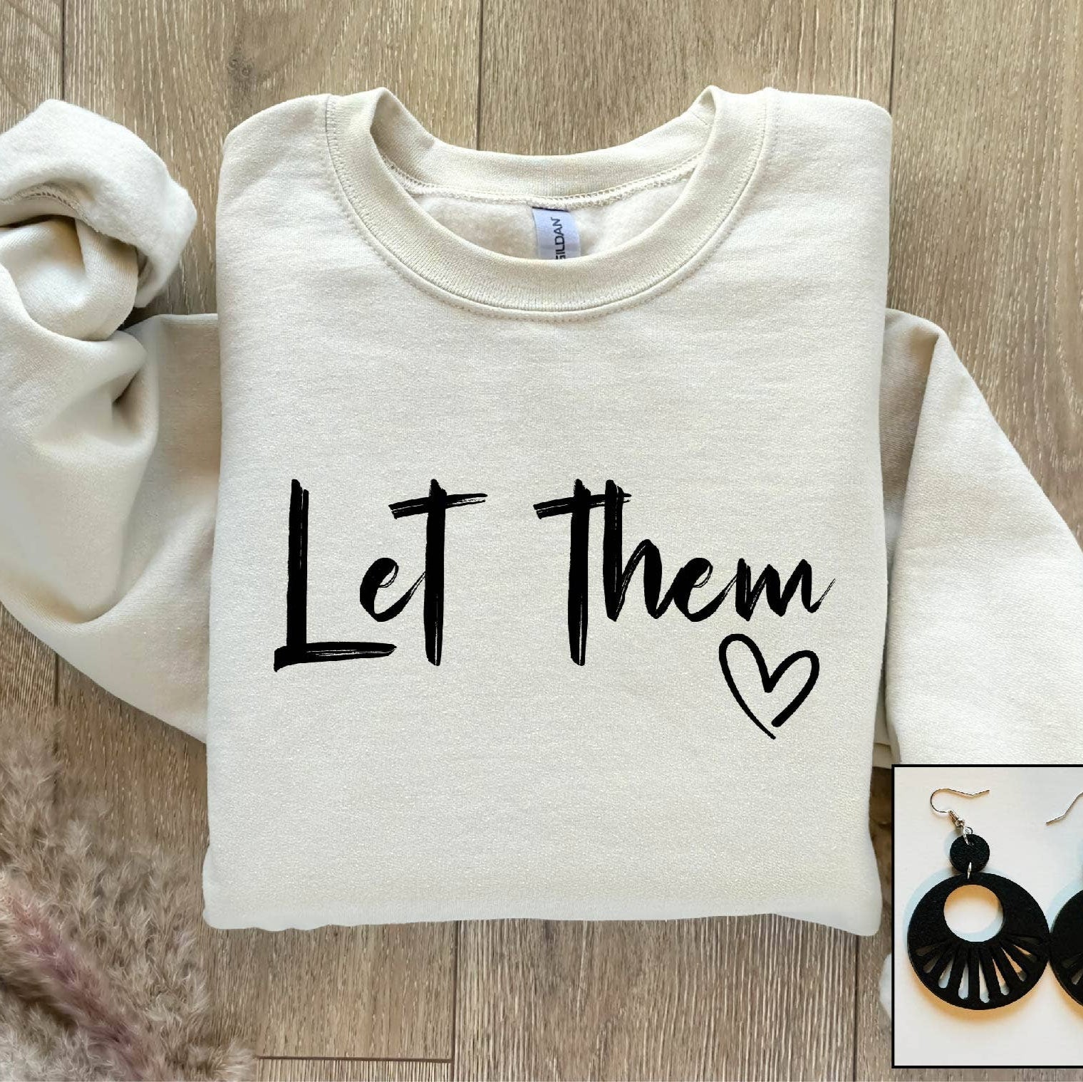 Folded white sweatshirt with 'Let Them' text and heart symbol on a wooden surface, with earrings displayed in an inset.