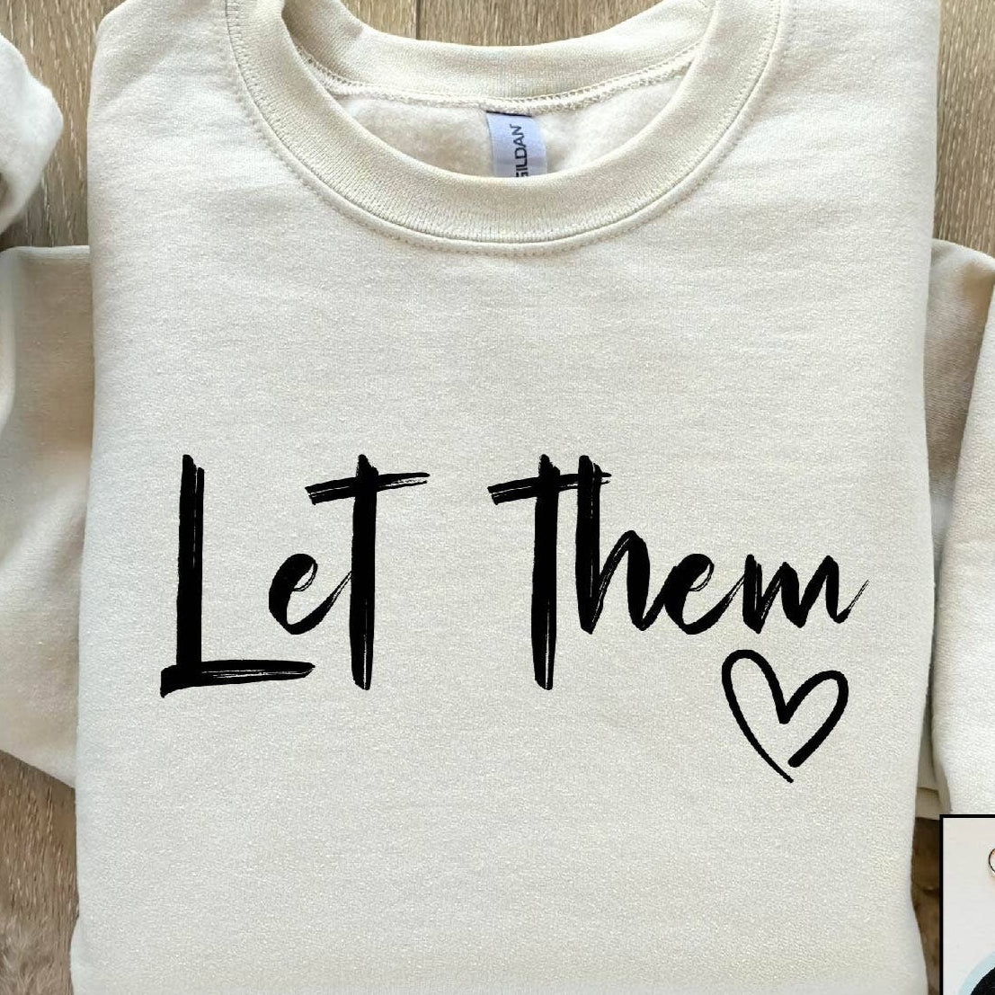 Folded white sweatshirt with 'Let Them' text and heart symbol on a wooden surface, with earrings displayed in an inset.