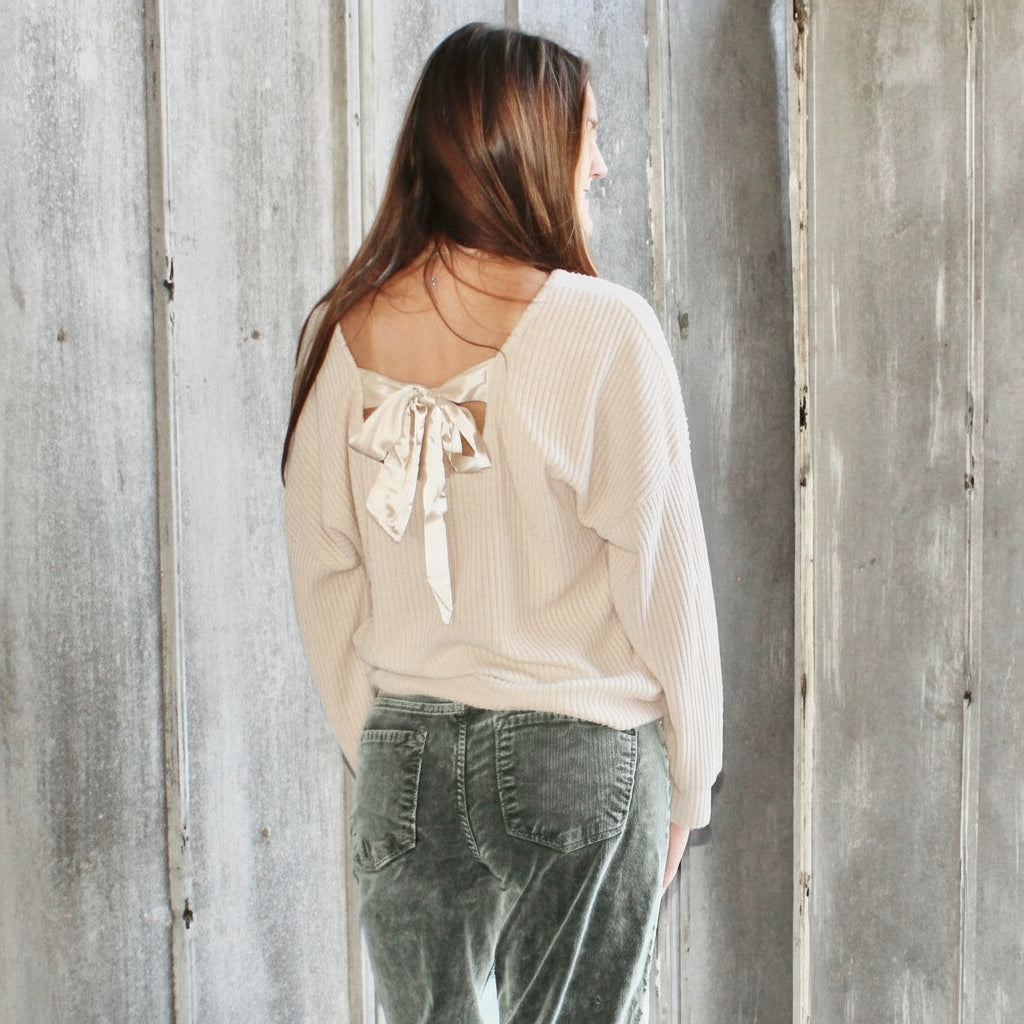 Woman wearing a light-colored blouse with a bow detail and dark pants, standing against a rustic wooden wall.
