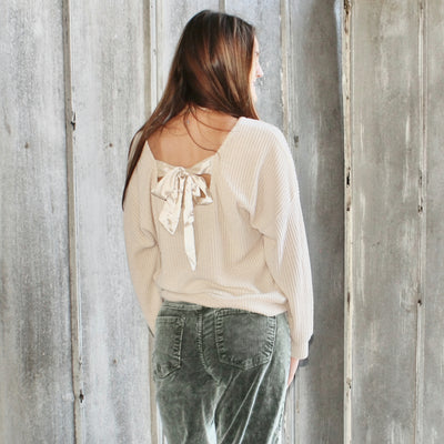 Woman wearing a light-colored blouse with a bow detail and dark pants, standing against a rustic wooden wall.