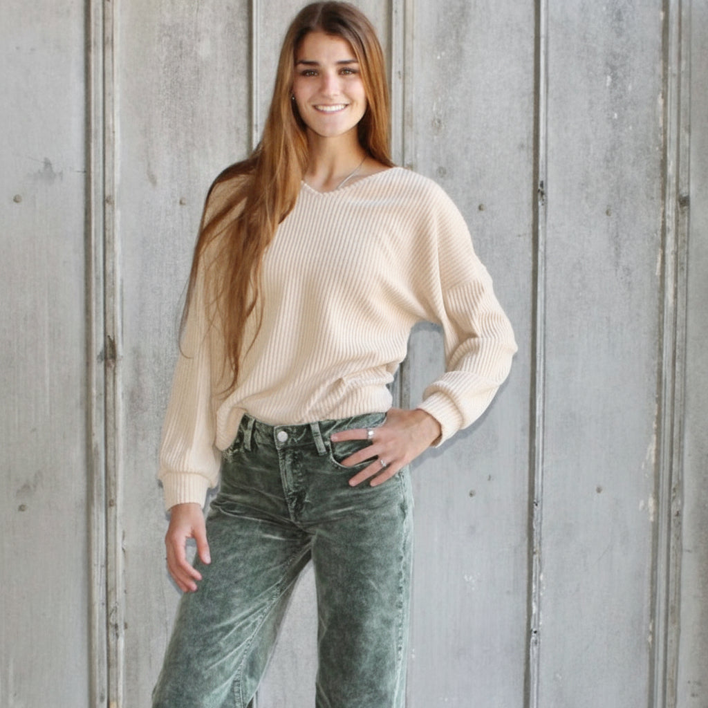 Woman wearing a beige sweater and green jeans standing against a wooden wall.