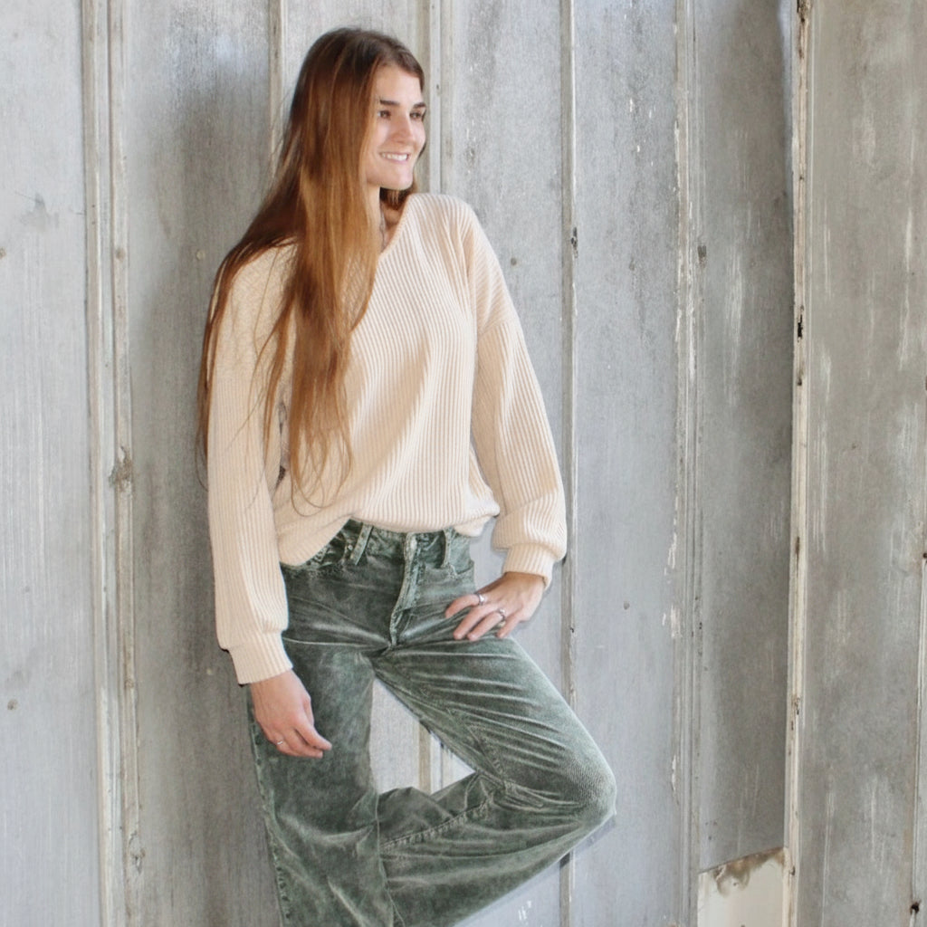 Woman wearing a beige sweater and green jeans standing against a wooden wall.
