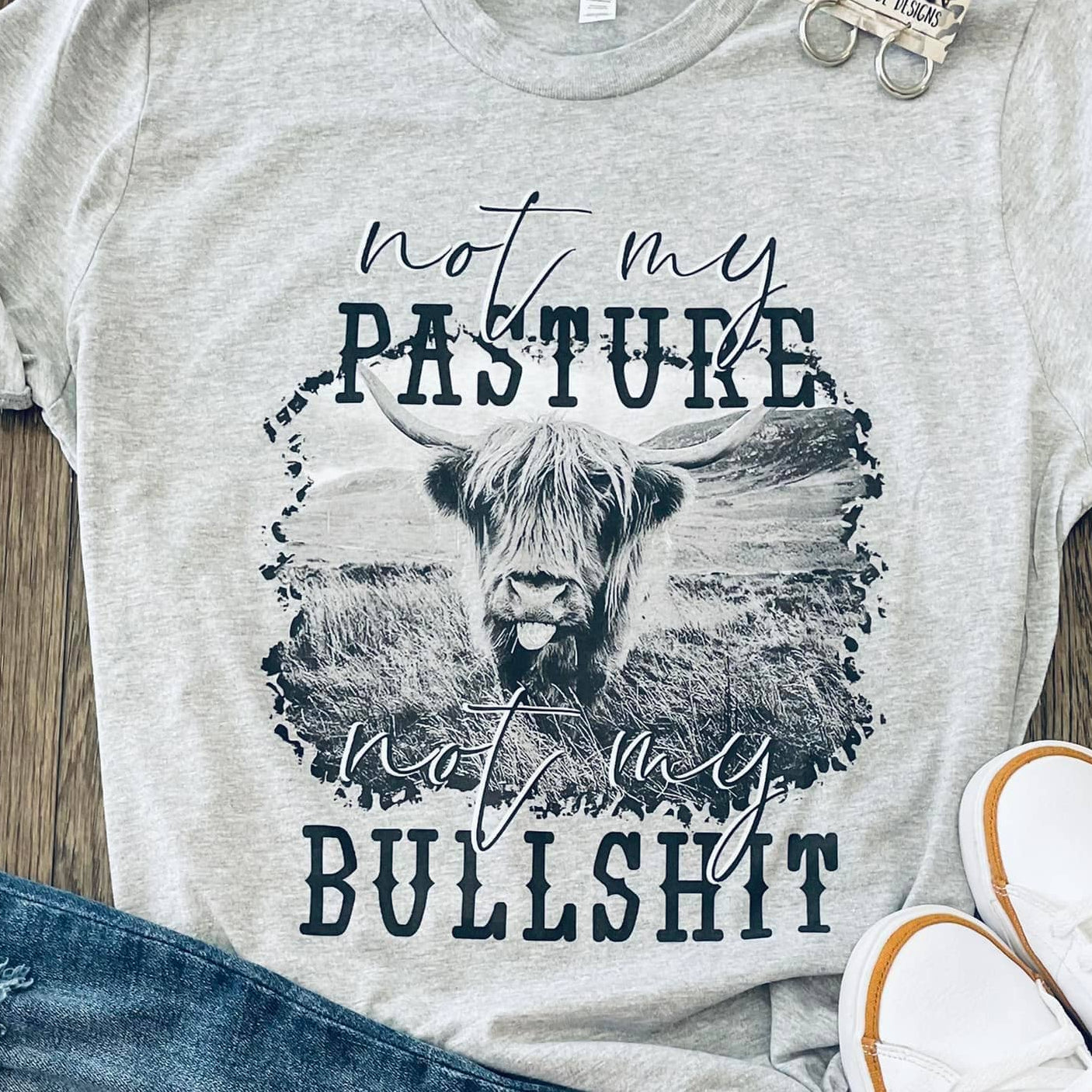 Gray t-shirt with graphic design and text, paired with jeans and sneakers on a wooden surface.