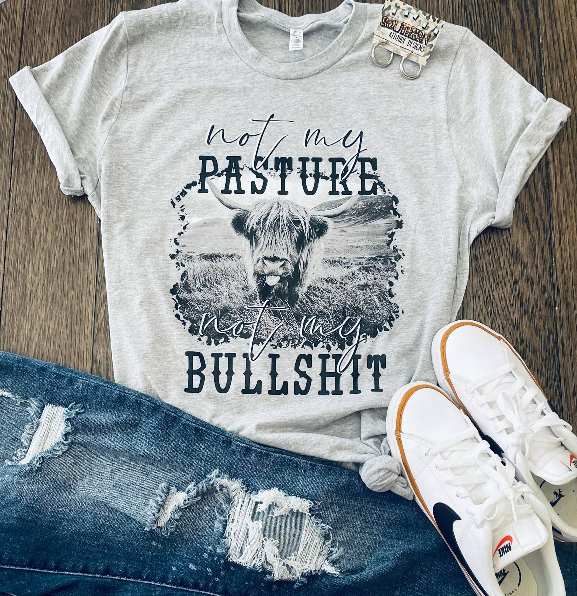 Gray t-shirt with graphic design and text, paired with jeans and sneakers on a wooden surface.