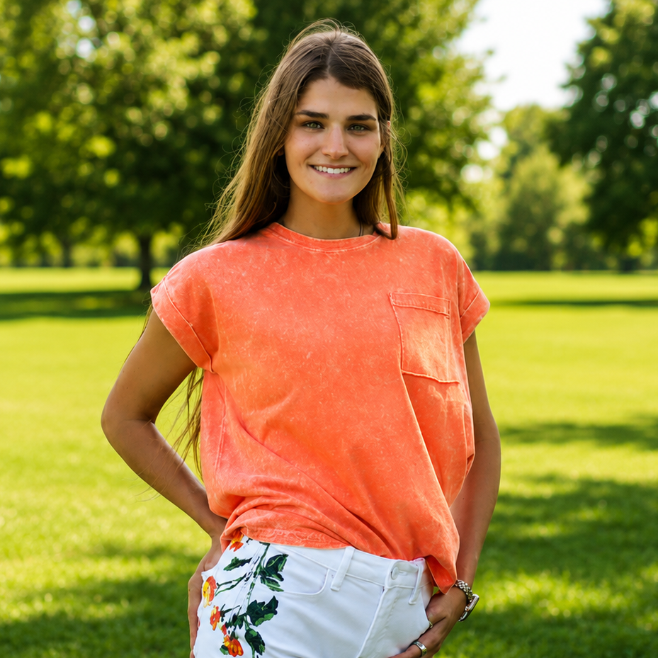 Woman wearing an orange t-shirt and white shorts with floral designs, standing in a grassy field.