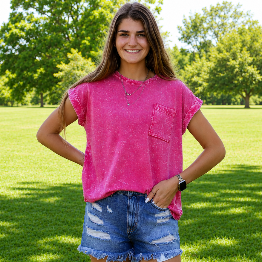 Woman in a pink shirt and denim shorts standing on grass with trees in the background