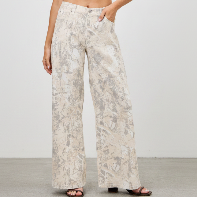 A woman modeling a pair of wide-leg pants with a beige and gray abstract pattern, paired with a light lavender crop top and brown sandals, against a plain light gray studio background.