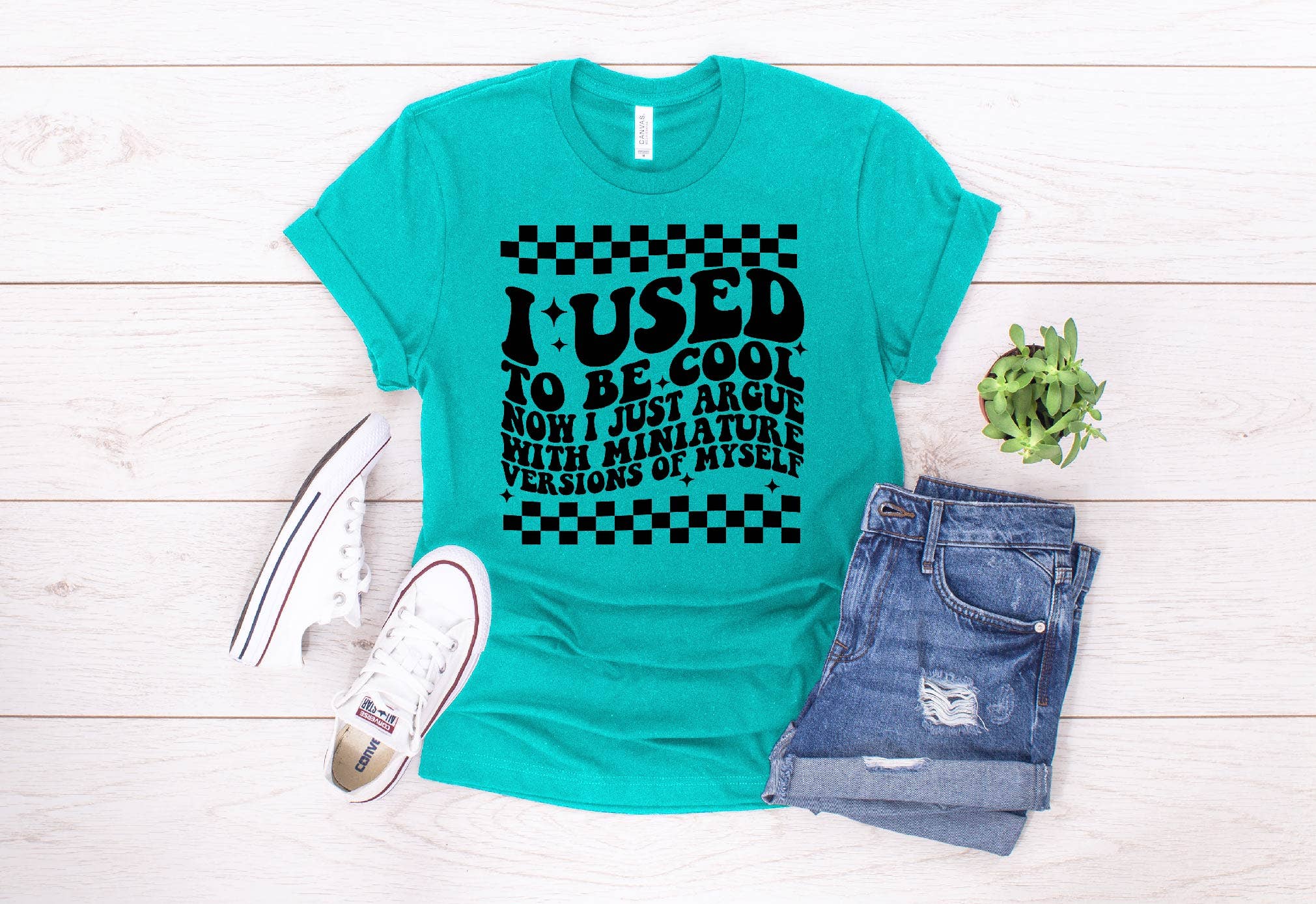 Turquoise t-shirt with humorous text design on a wooden surface with jeans and sneakers.