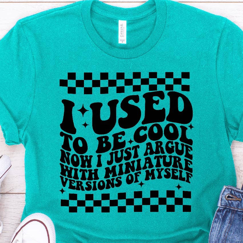 Turquoise t-shirt with humorous text design on a wooden surface with jeans and sneakers.