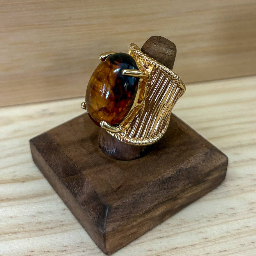 A gold ring with a large oval amber stone, displayed on a dark wooden stand over a light wood surface.