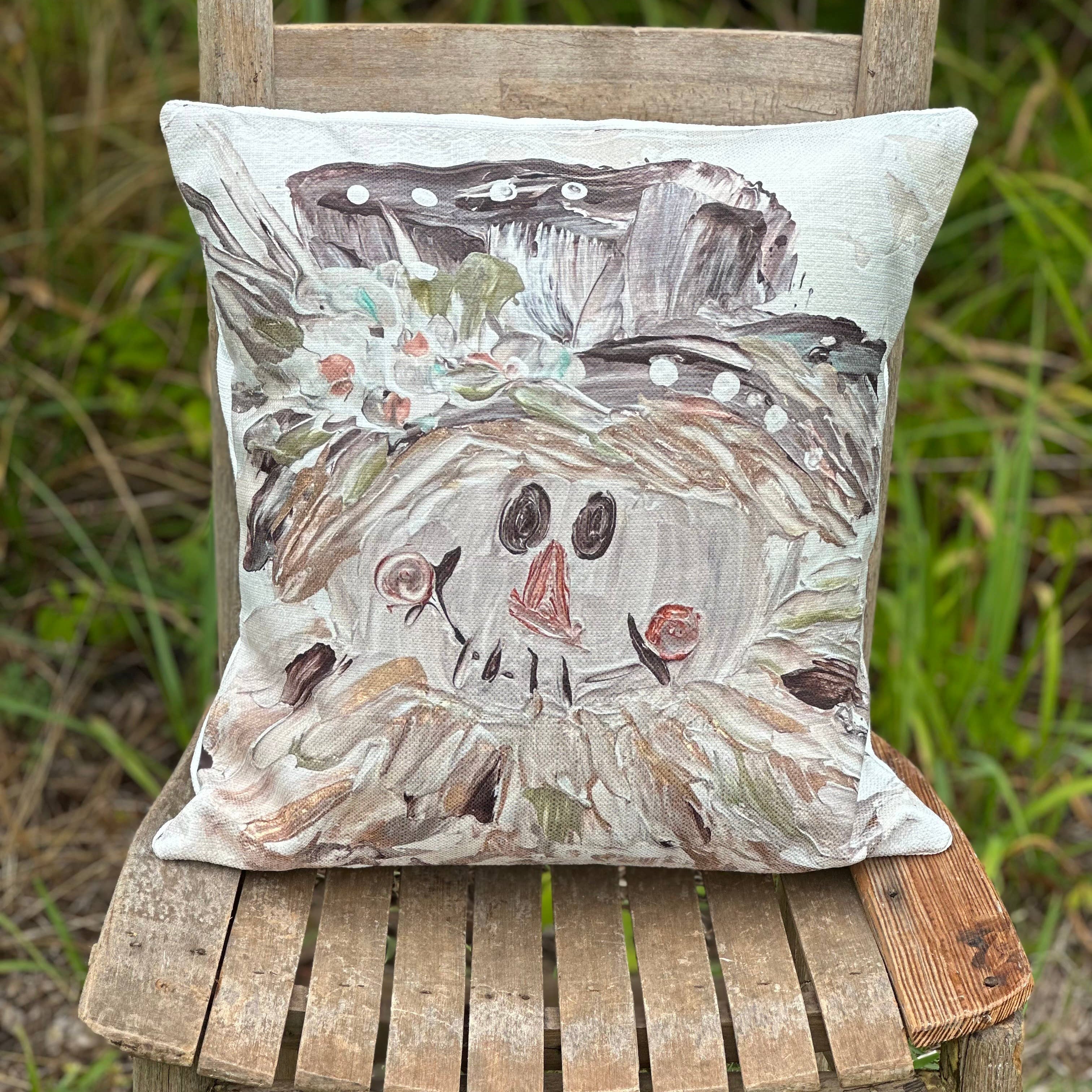 A square decorative throw pillow featuring a whimsical, hand-painted style snowman face and hat design, displayed on a rustic wooden chair in an outdoor garden setting.