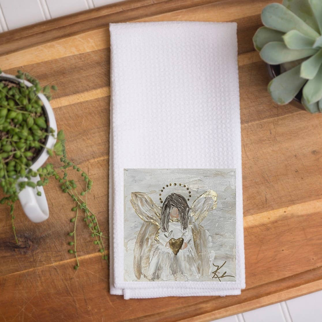 A folded white kitchen towel with a printed illustration of a winged angel, styled on a wooden cutting board with potted succulents.