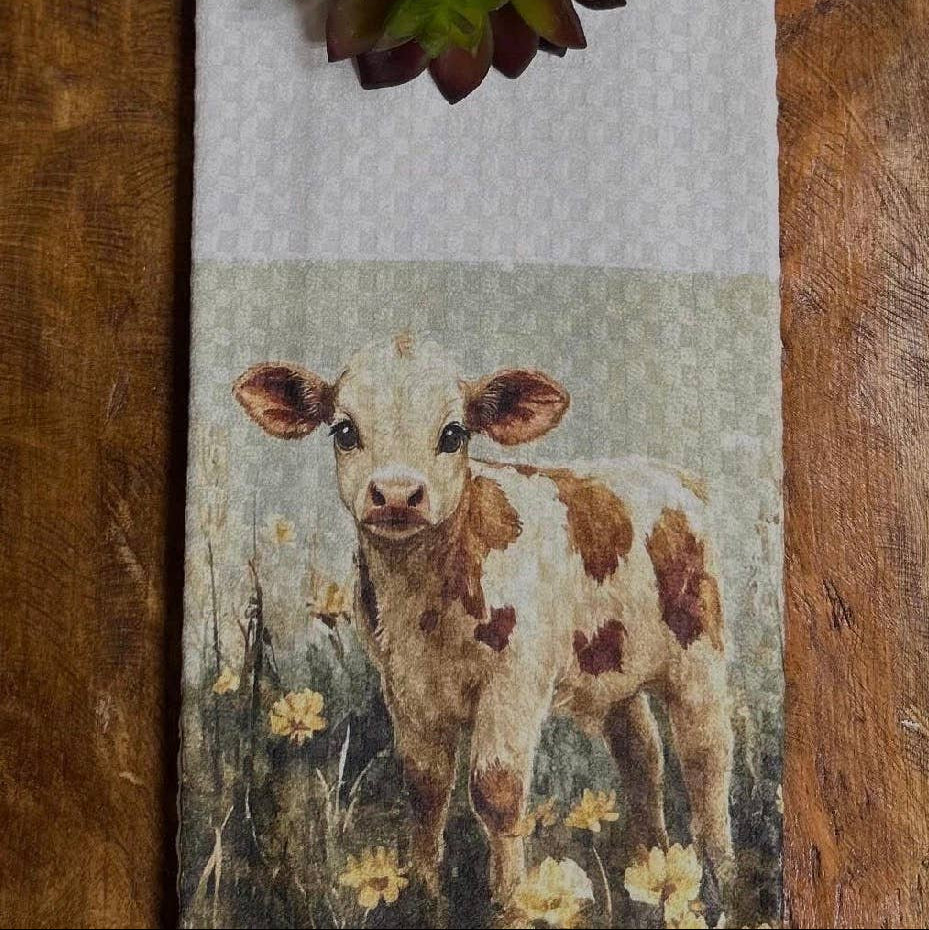 A rustic farmhouse-style woven table runner featuring a cow calf standing in a field of yellow flowers, styled on a dark wood table with a green succulent at the top.